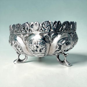 Puede incluir: Un cuenco de metal plateado con intrincados diseños en relieve de árboles, ciervos y otros animales. El borde está decorado con un patrón de peces repetido y el cuenco se apoya sobre tres patas con forma de pez, creando un recipiente metálico detallado.