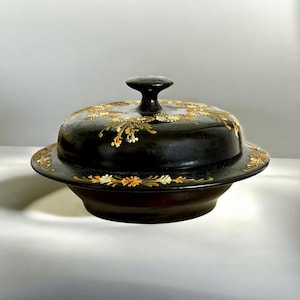 Puede incluir: Un plato negro con tapa, decorado con diseños florales dorados y naranjas. El plato tiene una base redonda y una tapa con un pequeño asa. El diseño floral adorna la tapa y el borde de la base.