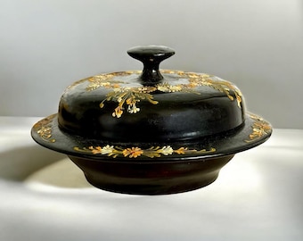 May include: A black, covered serving dish with a floral design in gold and orange. The dish has a round base and a lid with a small, rounded handle. The floral pattern decorates the lid and the rim of the base.