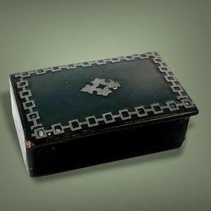 Puede incluir: Una pequeña caja rectangular azul oscuro con un diseño geométrico plateado en los bordes y un motivo floral central. La caja, con tapa, parece estar hecha de un material oscuro, posiblemente lacado, y es probablemente decorativa.