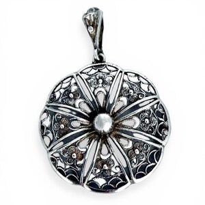 May include: A silver pendant with an intricate filigree design in a floral pattern. The pendant has a central, raised, circular element and a loop for a chain. The metal has a bright, reflective finish.