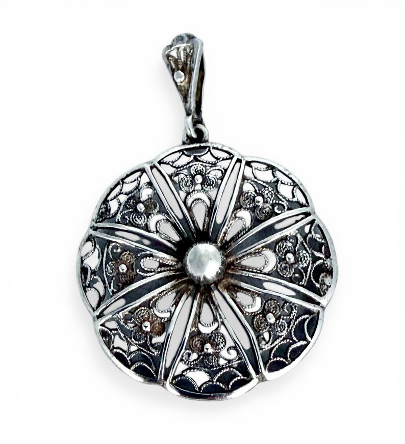 May include: A silver pendant with an intricate filigree design in a floral pattern. The pendant has a central, raised, circular element and a loop for a chain. The metal has a bright, reflective finish.