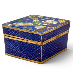 Puede incluir: Caja rectangular de esmalte azul con ribete dorado. Los lados presentan un patrón de escamas y la tapa un diseño floral con un jarrón y detalles dorados. Objeto decorativo.