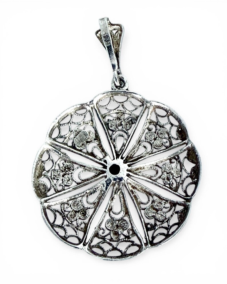May include: A silver filigree pendant with a floral design. The pendant features an openwork design with intricate details and small, sparkling accents. The pendant has a loop at the top for a chain and is set against a white background.