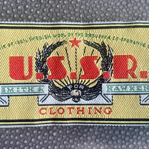 May include: A rectangular clothing label with the text "U.S.S.R." in red, framed by a black and gold design. The label also includes the text "SMITH & HAWKEN CLOTHING" and other text in green and black.