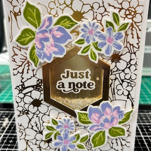 May include: A handmade greeting card with a floral design. The card features blue and pink flowers with green leaves, a gold hexagon with the text "Just a note", and a white background with a gold floral pattern.