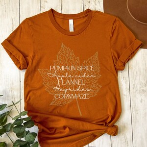 May include: A rust-colored t-shirt with a white graphic of a maple leaf with the text "Pumpkin Spice, Apple Cider, Flannel, Hayrides, Cornmaze".