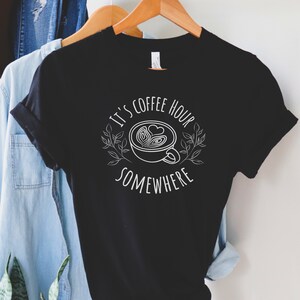 May include: Black t-shirt with white text that reads "It's coffee hour somewhere" and a stylized image of a coffee cup with a heart design.
