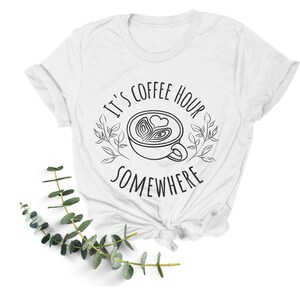 May include: White t-shirt with black text that reads "It's coffee hour somewhere" with a stylized drawing of a coffee cup with a heart design.