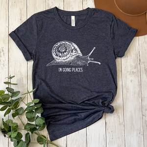 May include: A dark heather grey t-shirt with a white graphic of a snail and the text "I'm going places".