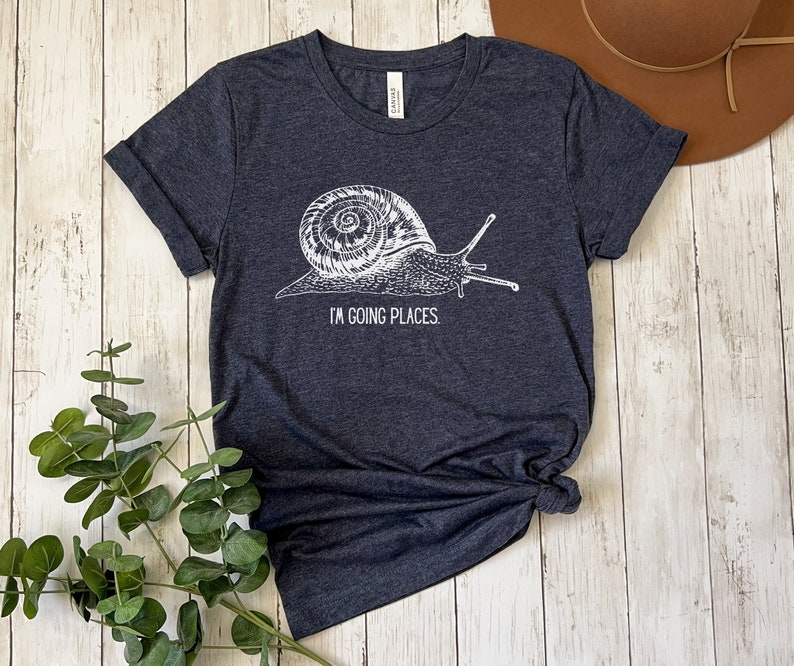 May include: A dark heather grey t-shirt with a white graphic of a snail and the text "I'm going places".