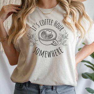 May include: A white t-shirt with the text "It's Coffee Hour Somewhere" and a stylized drawing of a coffee cup with a heart design.