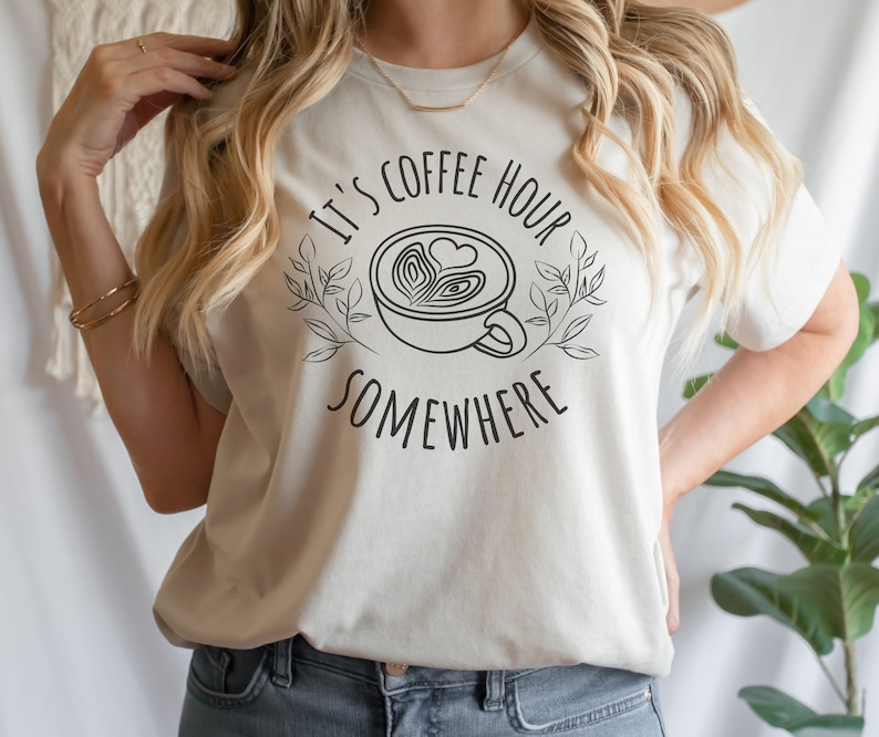 May include: A white t-shirt with the text "It's Coffee Hour Somewhere" and a stylized drawing of a coffee cup with a heart design.