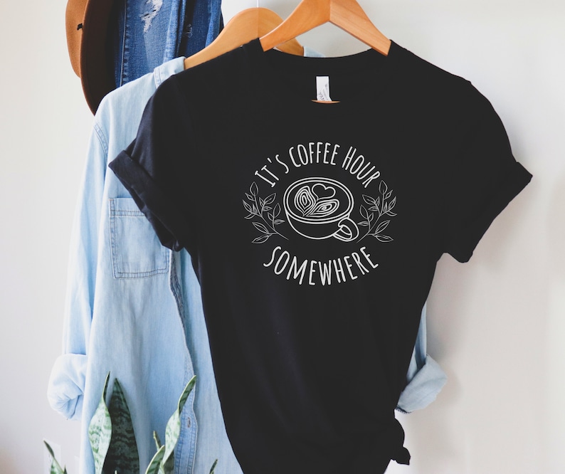 May include: Black t-shirt with white text that reads "It's coffee hour somewhere" and a stylized image of a coffee cup with a heart design.