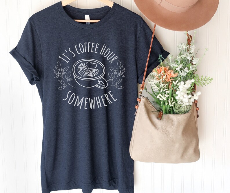 May include: A navy blue t-shirt with white text that reads "It's coffee hour somewhere" and a graphic of a coffee cup with a heart design.