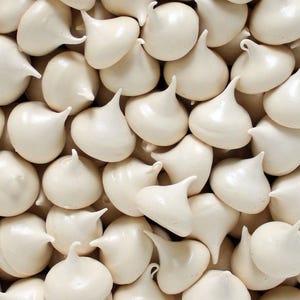 May include: A collection of numerous small, off-white meringue drops, each featuring a smooth, rounded base and a distinct, pointed peak. The individual pieces are tightly packed together, creating a uniform, textured surface of light cream-colored confections.