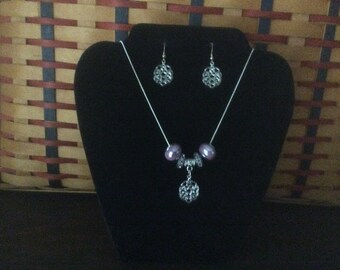 May include: A silver necklace with a silver pendant and two purple beads. The necklace is on a black display stand. The pendant is a silver filigree design. The earrings are silver filigree designs.