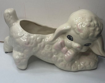 May include: White ceramic planter shaped like a lamb with pink accents and black eyes. The lamb is lying down with its head turned to the side. The planter has a hole in the top for drainage.