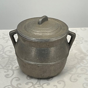 May include: A vintage, silver-colored, metal, lidded, round, storage container with two handles. The container has a smooth, slightly textured surface.
