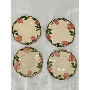 May include: Four cream-colored plates with scalloped edges. Each plate features a floral design with pink roses and green leaves around the rim. The rims have a gold-colored trim.