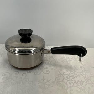 May include: A stainless steel saucepan with a lid and a black handle. The lid has a black knob. The handle is attached to the side of the pan. The pan is on a white surface.