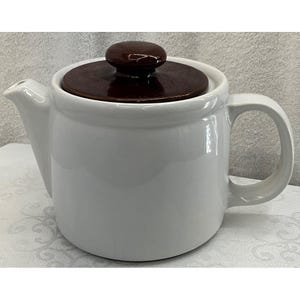 Op de afbeelding: Een witte keramische theepot met een bruin deksel en handvat. De theepot heeft een tuit om uit te schenken en een afgeronde body. Het deksel heeft een donkerbruine knop. De theepot is ontworpen voor het zetten van thee.