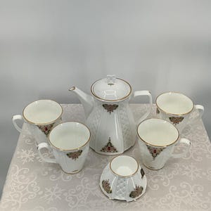 Puede incluir: Un juego de té de porcelana blanca con una tetera y seis tazas. El juego presenta ribetes dorados y un patrón decorativo con detalles en rojo y dorado. La tetera tiene tapa y un pico curvo. Las tazas tienen asas.