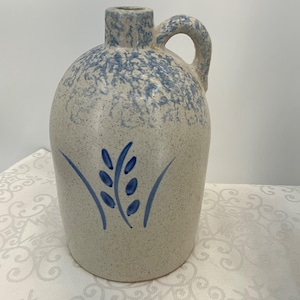 May include: A stoneware jug with a handle and a narrow neck. The jug is off-white with blue speckles on the top and a blue wheat stalk design on the front. The jug is a classic piece of kitchen decor.