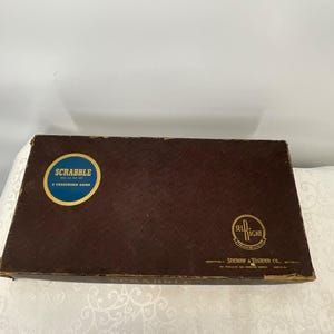 May include: A vintage Scrabble game box. The box is brown with a textured surface and features a circular label with the word "SCRABBLE" and the phrase "A CROSSWORD GAME". The Sel-Right logo is also visible.