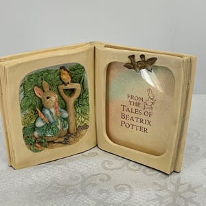 Puede incluir: Marco en forma de libro abierto de color crema con un relieve de Peter Rabbit y un pájaro. El lado izquierdo presenta a Peter Rabbit sosteniendo zanahorias, y el lado derecho tiene un marco de fotos con el texto: "From the Tales of Beatrix Potter."
