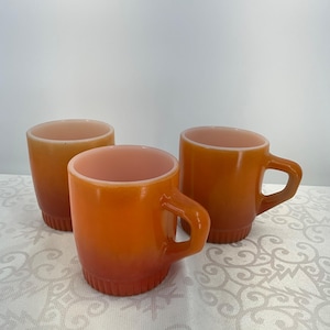 Puede incluir: Tres tazas vintage de vidrio naranja con efecto degradado. Las tazas tienen forma redondeada con asa y base acanalada. El interior de las tazas es de color rosa claro.