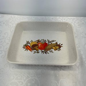 May include: Rectangular off-white ceramic baking dish with a speckled finish. The centre features a colourful illustration of vegetables including carrots, tomatoes, corn, and mushrooms. The dish has a slightly raised, ridged edge.
