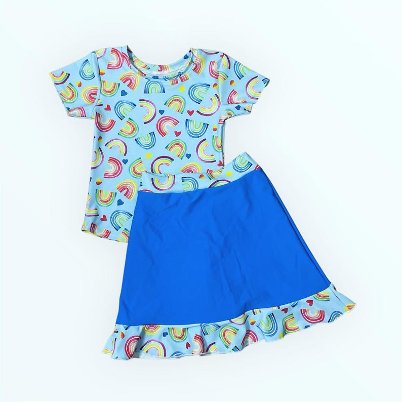 Puede incluir: Un conjunto de dos piezas para ni&ntilde;as que incluye una camiseta de manga corta y una falda azul. La camiseta tiene un fondo azul claro con un estampado de arco&iacute;ris y corazones. La falda es azul con un ribete de volantes de arco&iacute;ris y corazones.