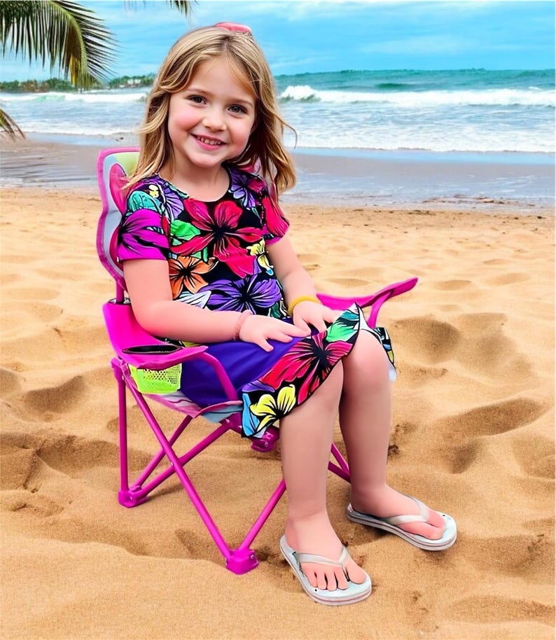 Puede incluir: Una ni&ntilde;a peque&ntilde;a se sienta en una silla plegable rosa con forma de mariposa en una playa de arena. La silla tiene un portavasos de malla y un estampado floral. La ni&ntilde;a lleva un vestido floral y chanclas.