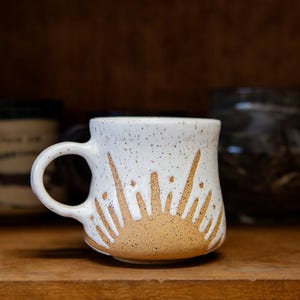 May include: A white ceramic mug with a speckled finish and a tan sunburst design at the base. The mug features a rounded handle and a slightly flared rim, showcasing a hand-crafted appearance with visible clay texture and a speckled glaze pattern.