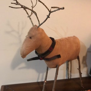 May include: A rustic, tan-colored reindeer figurine with twig antlers and legs. The reindeer has a stitched face and a plaid scarf around its neck. This decorative item is likely handmade and suitable for holiday or seasonal decor.