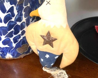 Può includere: Un ornamento di aquila patriottico fatto a mano con corpo giallo, ali blu con stelle bianche e una stella marrone sul petto. L'aquila ha un becco giallo e una testa bianca con una X nera per gli occhi.