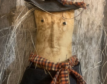 May include: A handmade scarecrow doll with a tan face, black hat, and black outfit. The doll has a plaid scarf in orange and brown. Wisps of straw-like material extend from the head. The doll is a rustic, decorative item.