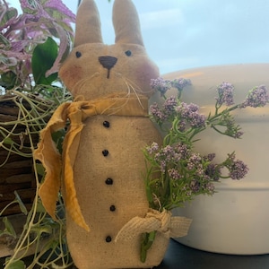 May include: A handmade burlap bunny with a yellow ribbon around its neck and a bouquet of purple flowers in its hand.