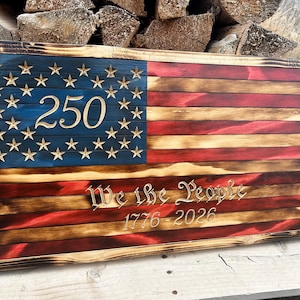 May include: A rectangular wooden wall sign featuring a charred, rustic American flag design. The blue canton displays the number 250, while the red and white stripes feature the engraved text We the People and the years 1776-2026 in a serif font.