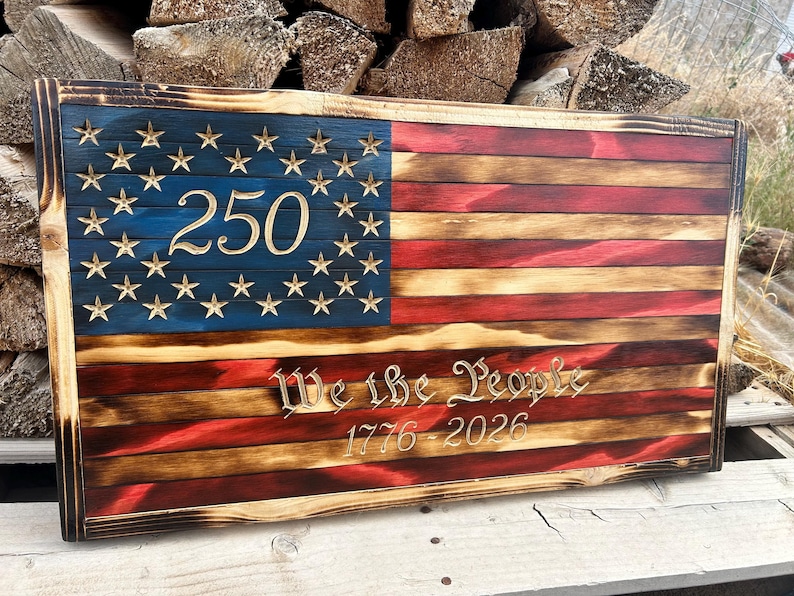 May include: A rectangular wooden wall sign featuring a charred, rustic American flag design. The blue canton displays the number 250, while the red and white stripes feature the engraved text We the People and the years 1776-2026 in a serif font.
