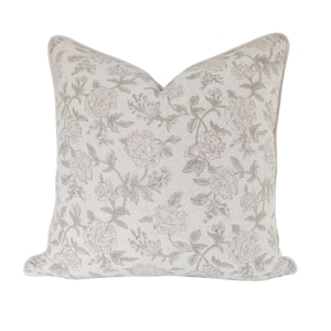 May include: Square decorative pillow with a floral pattern. The pillow features a cream-colored background with a repeating design of pink and gray flowers and leaves. The pillow has a soft, inviting appearance.