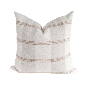 May include: A square throw pillow featuring a textured off-white fabric with vertical stripes in a light beige tone. The design includes horizontal bands of thin, frayed-edge brown lines that cross the vertical stripes across the front surface.