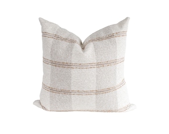 May include: A square throw pillow featuring a textured off-white fabric with vertical stripes in a light beige tone. The design includes horizontal bands of thin, frayed-edge brown lines that cross the vertical stripes across the front surface.