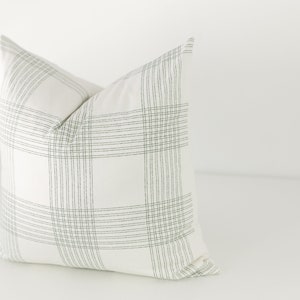 May include: A square throw pillow featuring a white base fabric with a dark green grid pattern. The design consists of sets of thin, parallel vertical and horizontal lines that intersect to form a large, repeating plaid-like check motif.