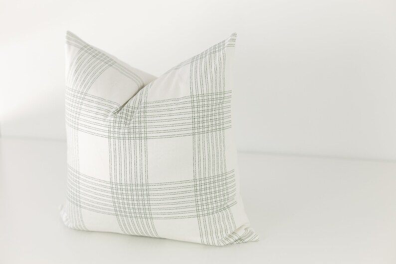 May include: A square throw pillow featuring a white base fabric with a dark green grid pattern. The design consists of sets of thin, parallel vertical and horizontal lines that intersect to form a large, repeating plaid-like check motif.