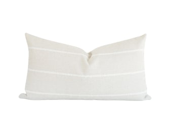 May include: A rectangular lumbar pillow featuring a light beige fabric base with three horizontal white stripes. The pillow has a soft, textured appearance with a slight indentation at the top center, creating a structured yet plush look.