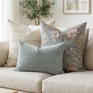 May include: A collection of decorative throw pillows in various patterns and colors. The pillows are arranged on a beige sofa. One pillow is a solid light blue, another has a floral design, and the third has a striped pattern.