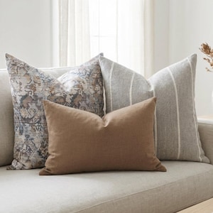 May include: A collection of decorative throw pillows in various patterns and colors. The pillows include a brown rectangular pillow, a gray striped pillow, and two patterned pillows with blue, brown, and beige tones. The pillows are arranged on a light-colored sofa.