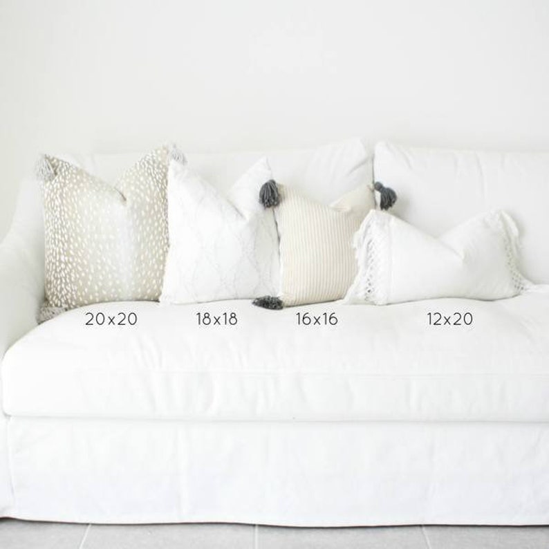 May include: A collection of decorative throw pillows in various sizes and textures, arranged on a white sofa. Pillows include a 20x20 inch spotted design, an 18x18 inch embroidered pattern, a 16x16 inch ribbed texture, and a 12x20 inch fringed pillow.